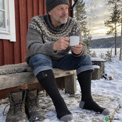Mies punaisen mökin edessä lumessa lämpösukat jalassa ja kahvikuppi kädessä
